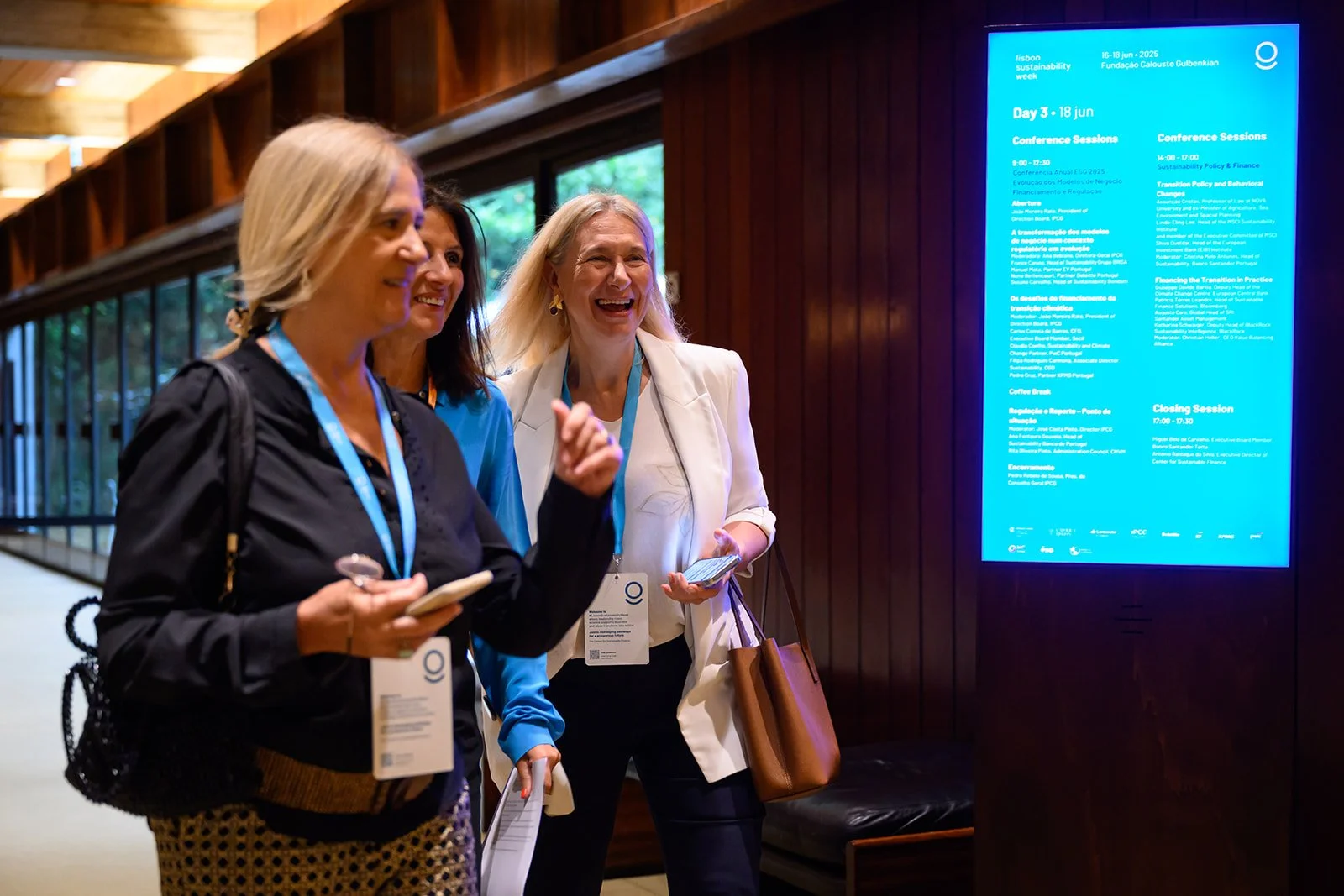 Three women at a conference looking at a large blue digital schedule board, smiling, and sharing a light moment.