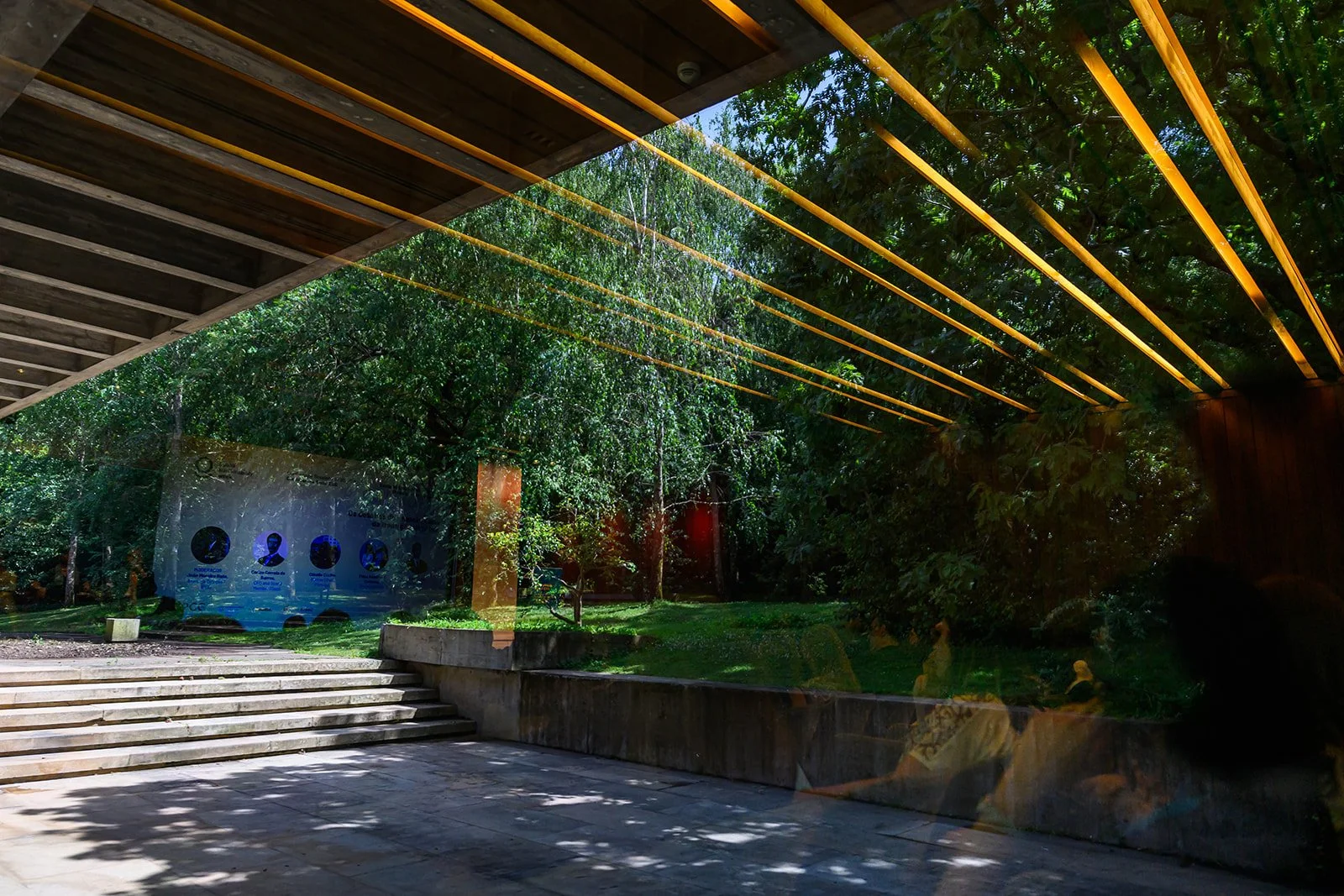 View through a glass window reflecting an outdoor scene with trees, a concrete staircase, and people sitting on benches.