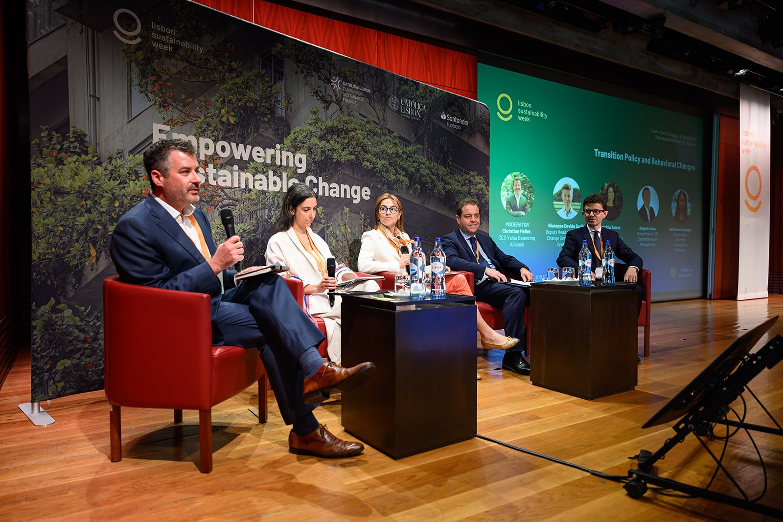 Panel discussion at Lisbon Sustainability Week with five professionals sitting on stage, microphones, and water bottles, backdrop shows event branding with a cityscape, and a projector screen displaying session details.