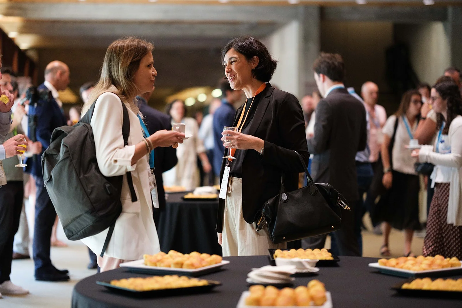 Two women conversing at a networking event with food trays and other attendees in the background.