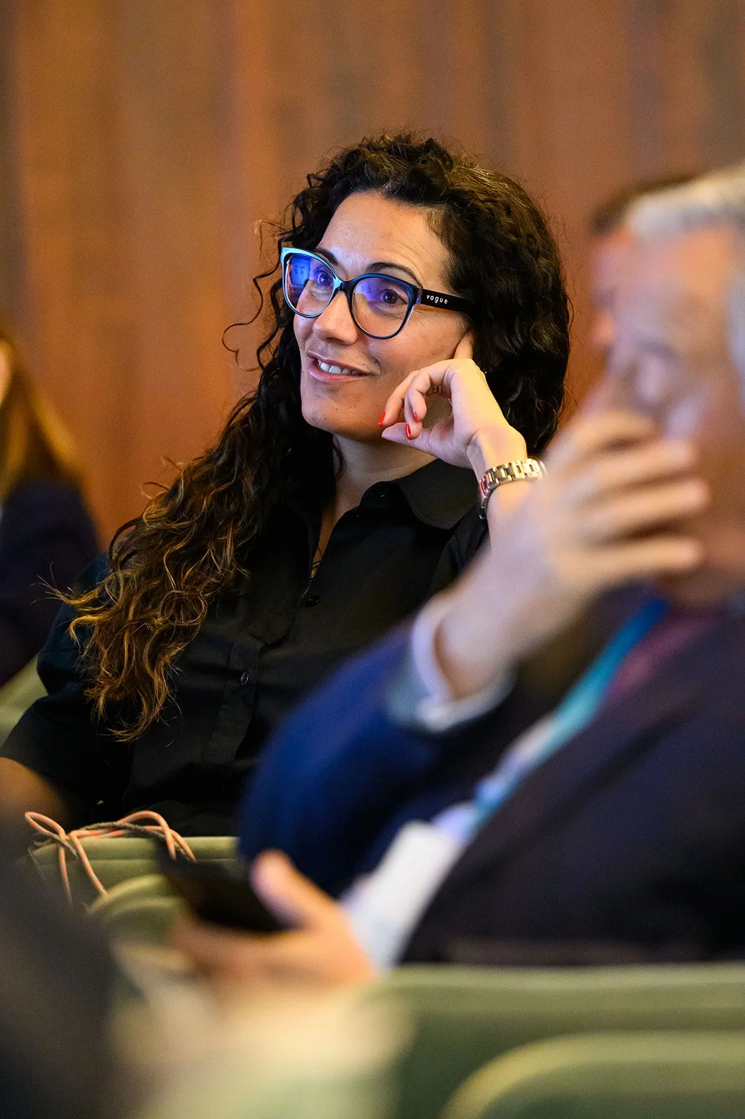 A woman with curly dark hair and glasses sitting in a conference room, smiling and listening attentively, with a man next to her looking down at a phone.