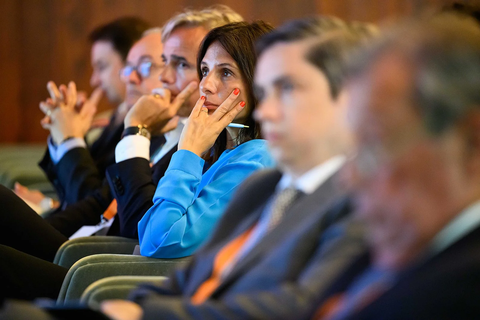 A group of five people in formal attire seated in an auditorium, appear to be paying attention to a presentation or speech. One woman in a blue shirt is in focus, resting her chin on her hand with a thoughtful expression.