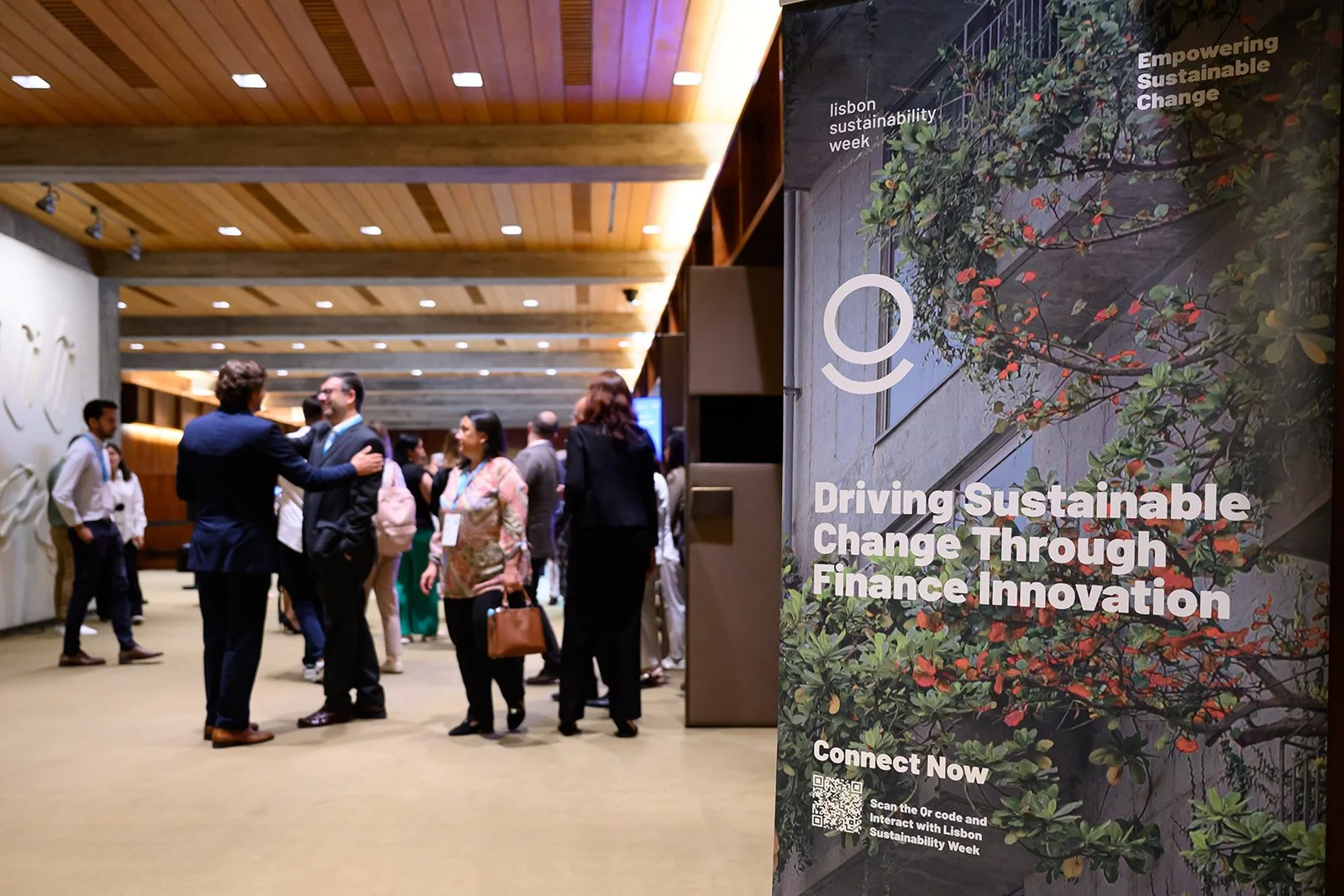 A group of people engaging in conversation at a conference or event inside a venue with wooden ceiling panels. A large sign on the right reads, 'Driving Sustainable Change Through Finance Innovation,' with mentions of Lisbon Sustainability Week and a