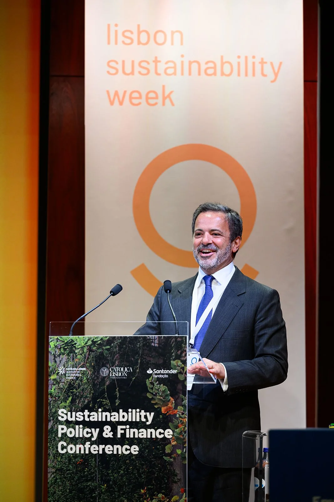 A man in a suit and tie standing at a podium labeled 'Sustainability Policy & Finance Conference,' speaking at Lisbon Sustainability Week.