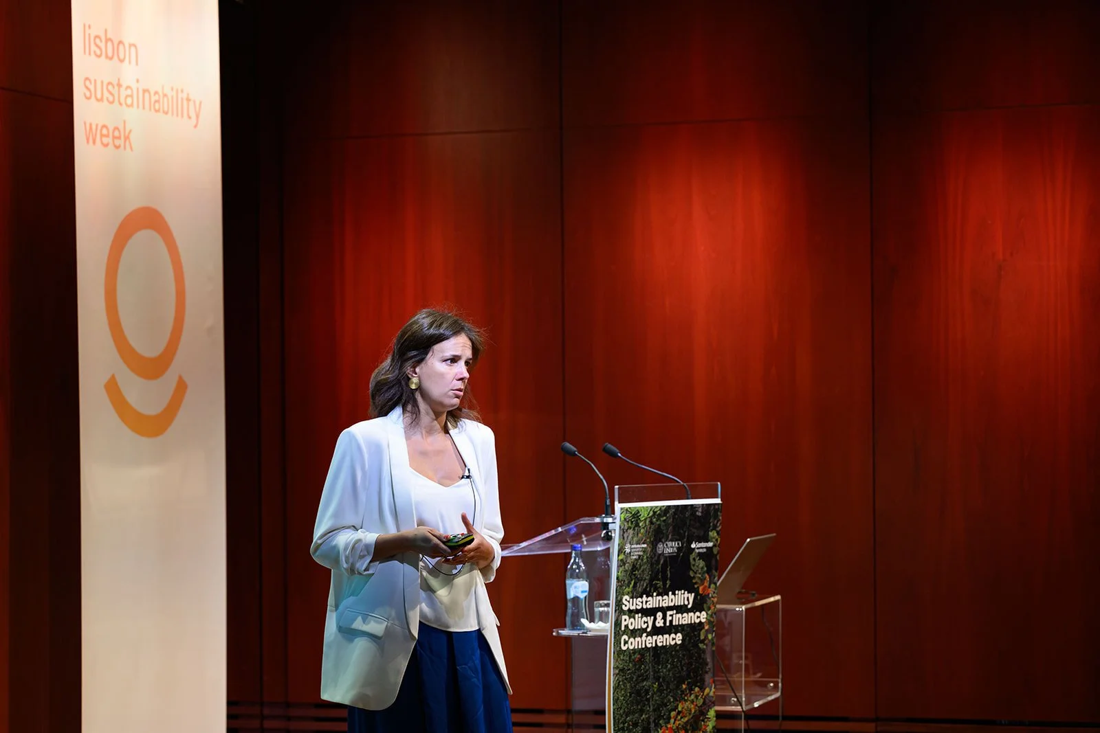 A woman in white blazer and navy skirt giving a presentation at a conference about sustainability policy and finance, with a banner that reads 'Lisbon Sustainability Week' and a lectern with a sign that says 'Sustainability Policy & Finance Conferenc
