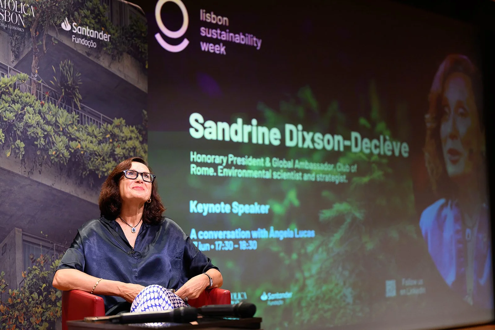 A woman with glasses, wearing a dark blouse and patterned pants, sitting in a red chair on stage at the Lisbon Sustainability Week event, with a large presentation screen behind her displaying her name, title, and details of a conversation with Ágath