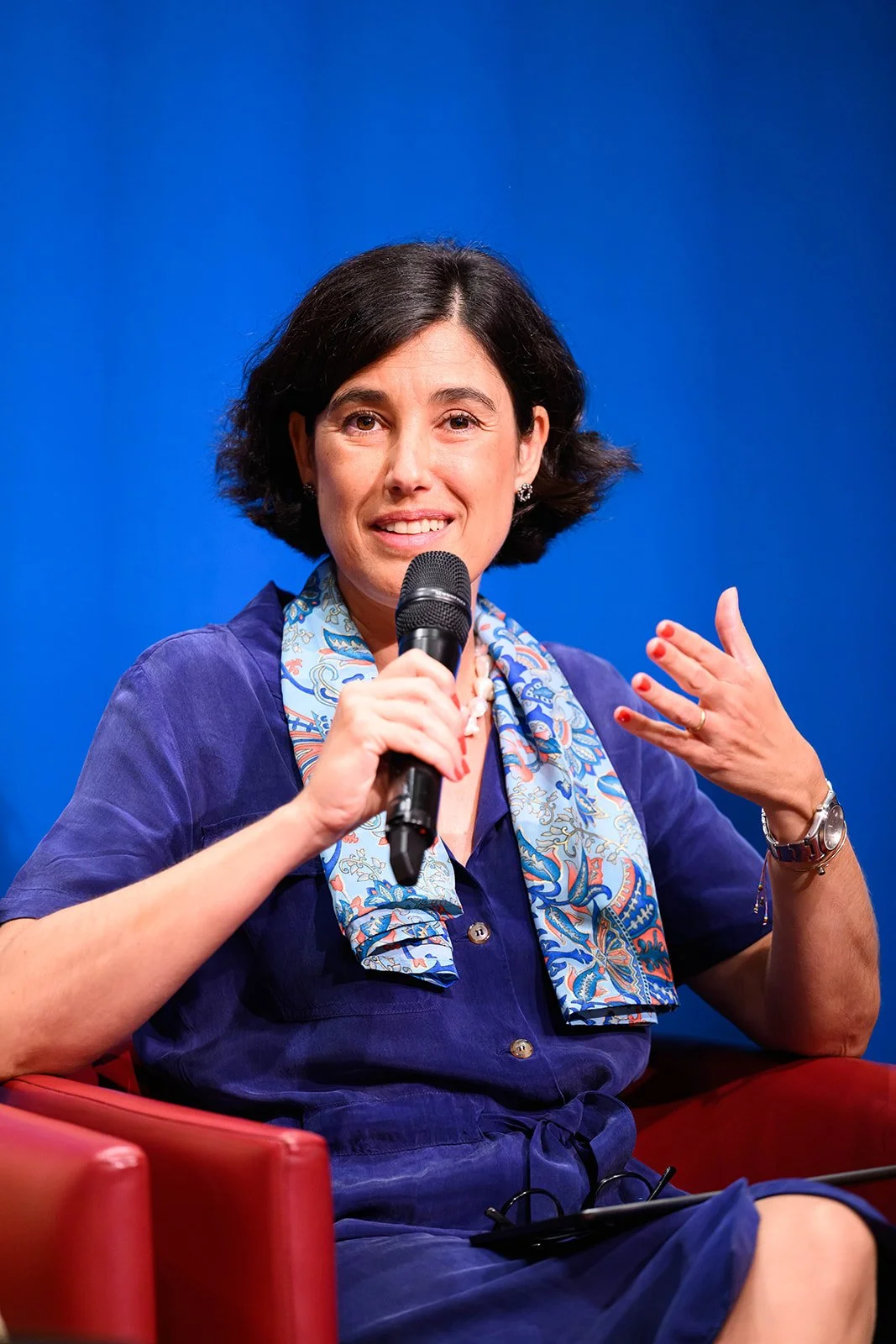 Woman with short dark hair holding a microphone and speaking, dressed in a navy blue outfit with a patterned scarf, sitting on a red chair against a blue background.