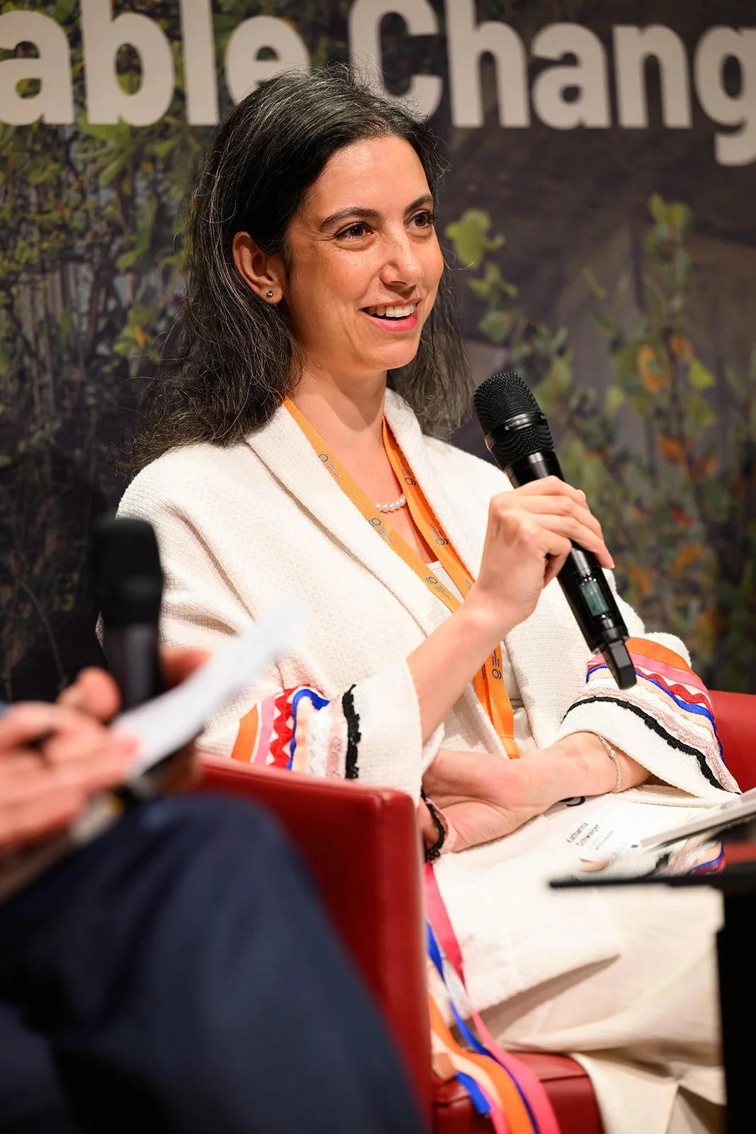 A woman with dark hair, wearing a white jacket with colorful embroidered patterns on the sleeves, holding a microphone and speaking at an event or panel. There is a background with text and some greenery.