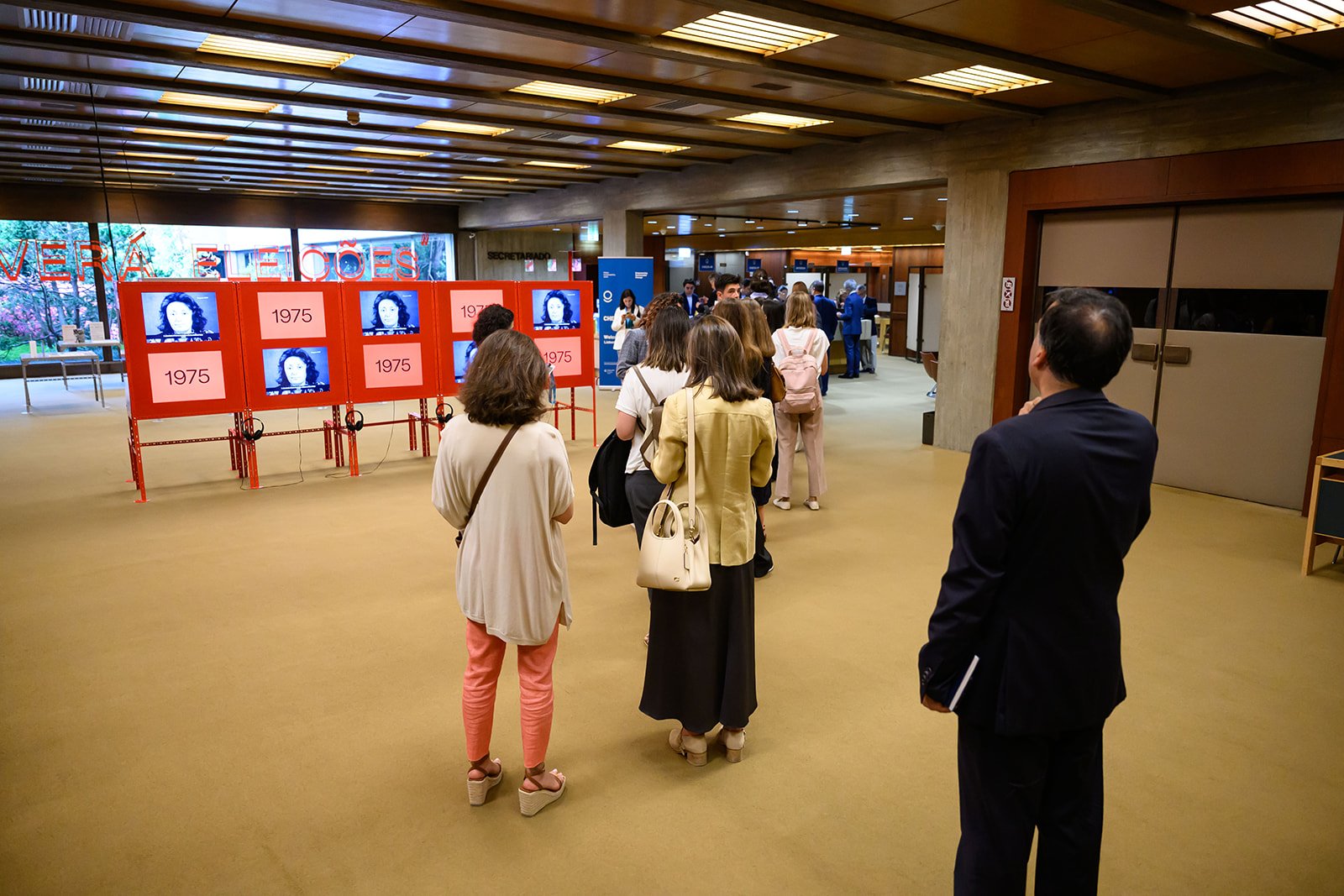 People waiting in line inside a building with red display boards showing television screens and the year 1975, during an event or exhibit.