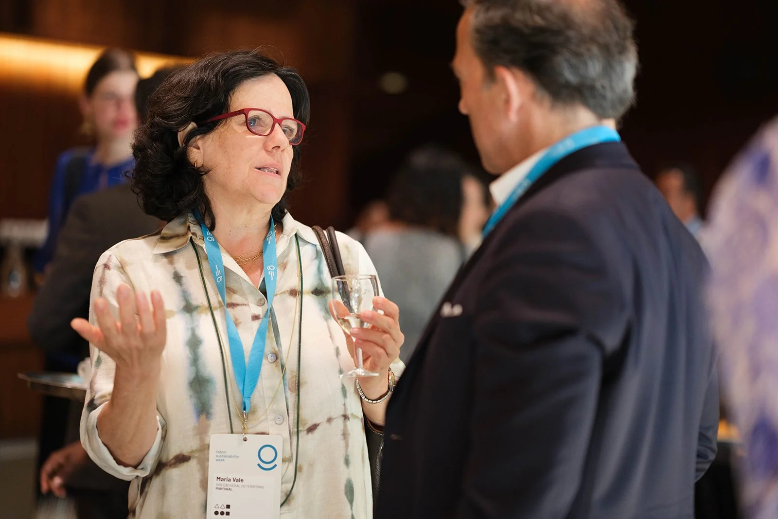 A woman with dark hair and red glasses talking to a man at a professional event, holding a wine glass, wearing identification badge.