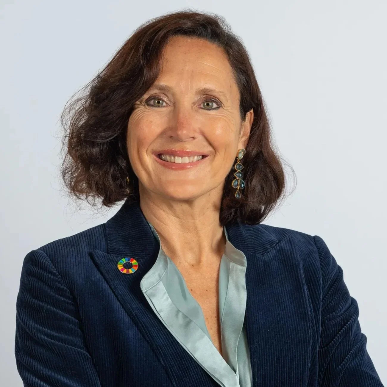 Smiling woman in blue blazer with a circular pin and decorative earrings.