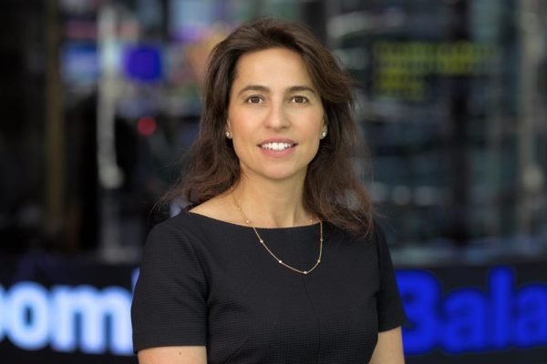 A woman with long brown hair wearing a black dress and a necklace, standing indoors with blurred background.