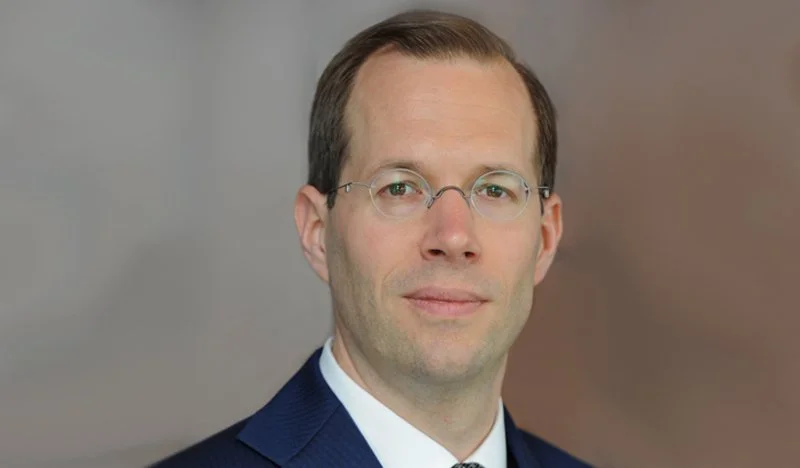 Man wearing glasses and a suit with a neutral expression, posing against a plain background.