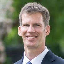 Man in a suit smiling outdoors with a blurred green background.