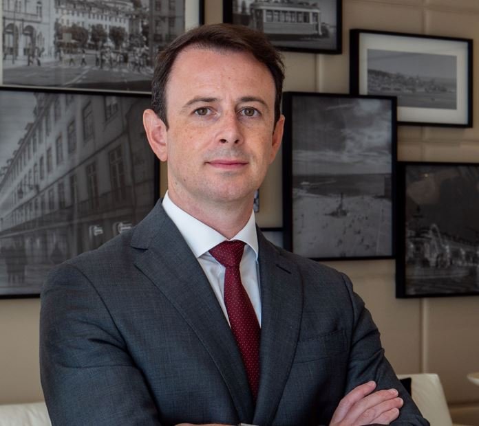 Man in a suit with a red tie standing in front of a wall with framed photographs.