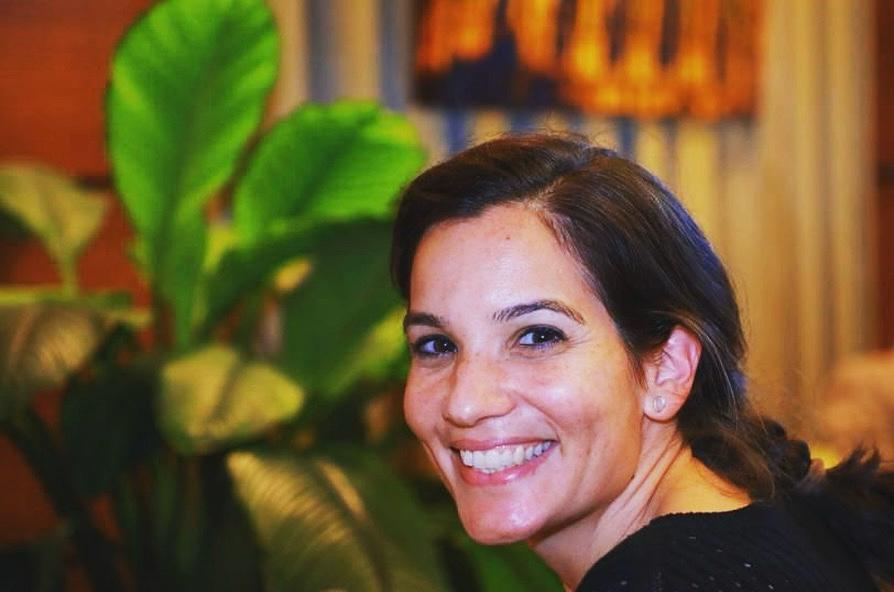 Smiling woman with dark hair sits in front of a leafy green plant indoors.