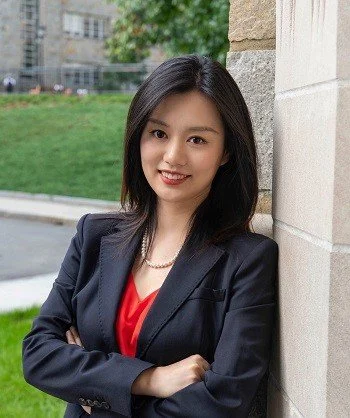 Businesswoman standing outdoors with arms crossed, wearing a black blazer and red blouse, smiling at the camera.