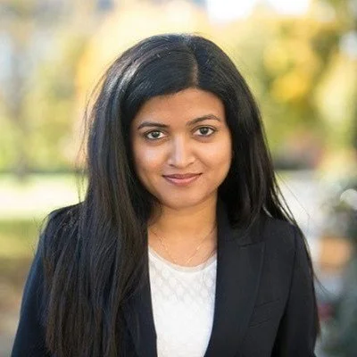 Portrait of a person with long dark hair in an outdoor setting, wearing a dark blazer and light blouse.