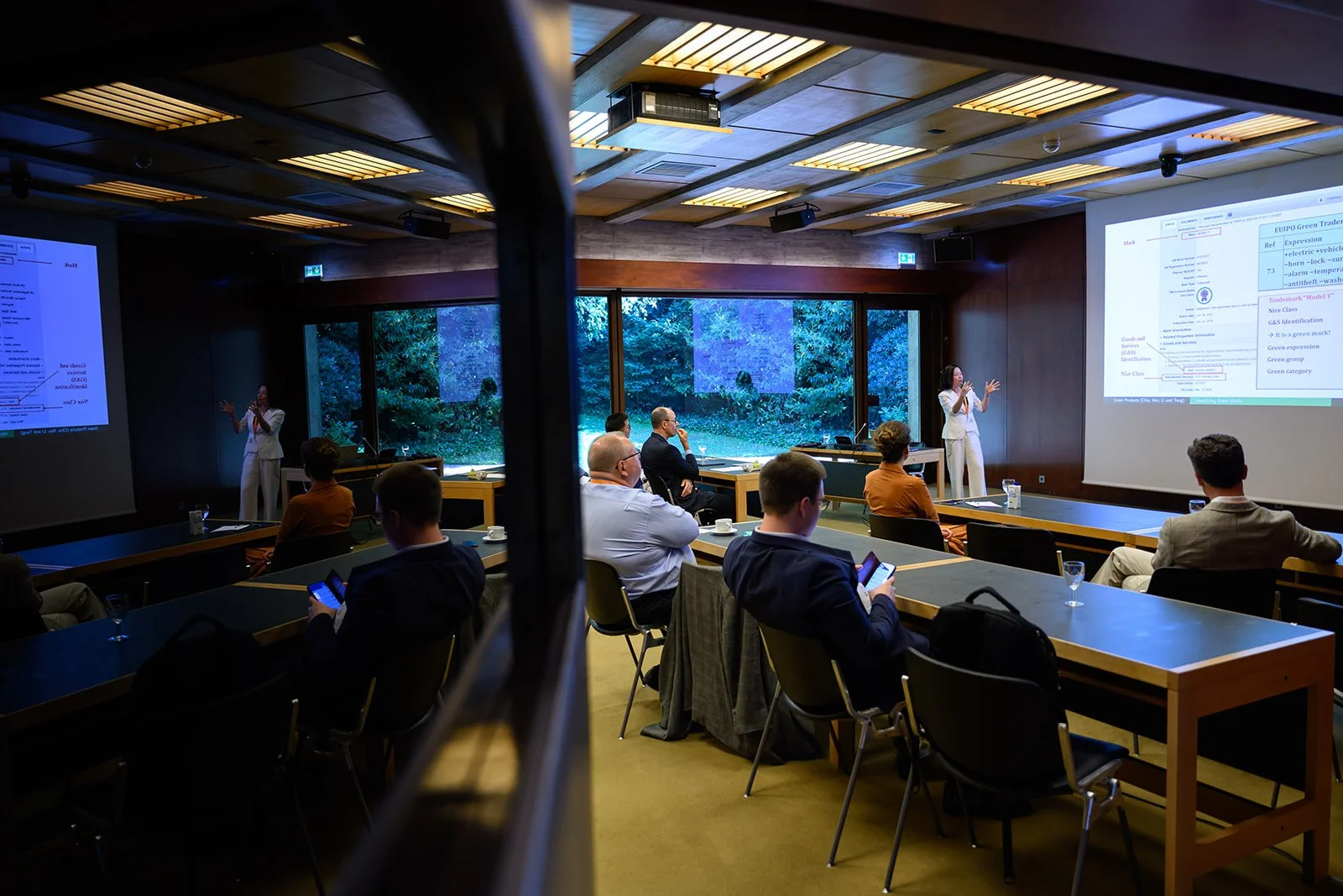 A woman is giving a presentation in a conference room with several attendees seated at tables, some using their phones. Two large screens display slides relevant to the presentation. The room has large windows showing trees outside.