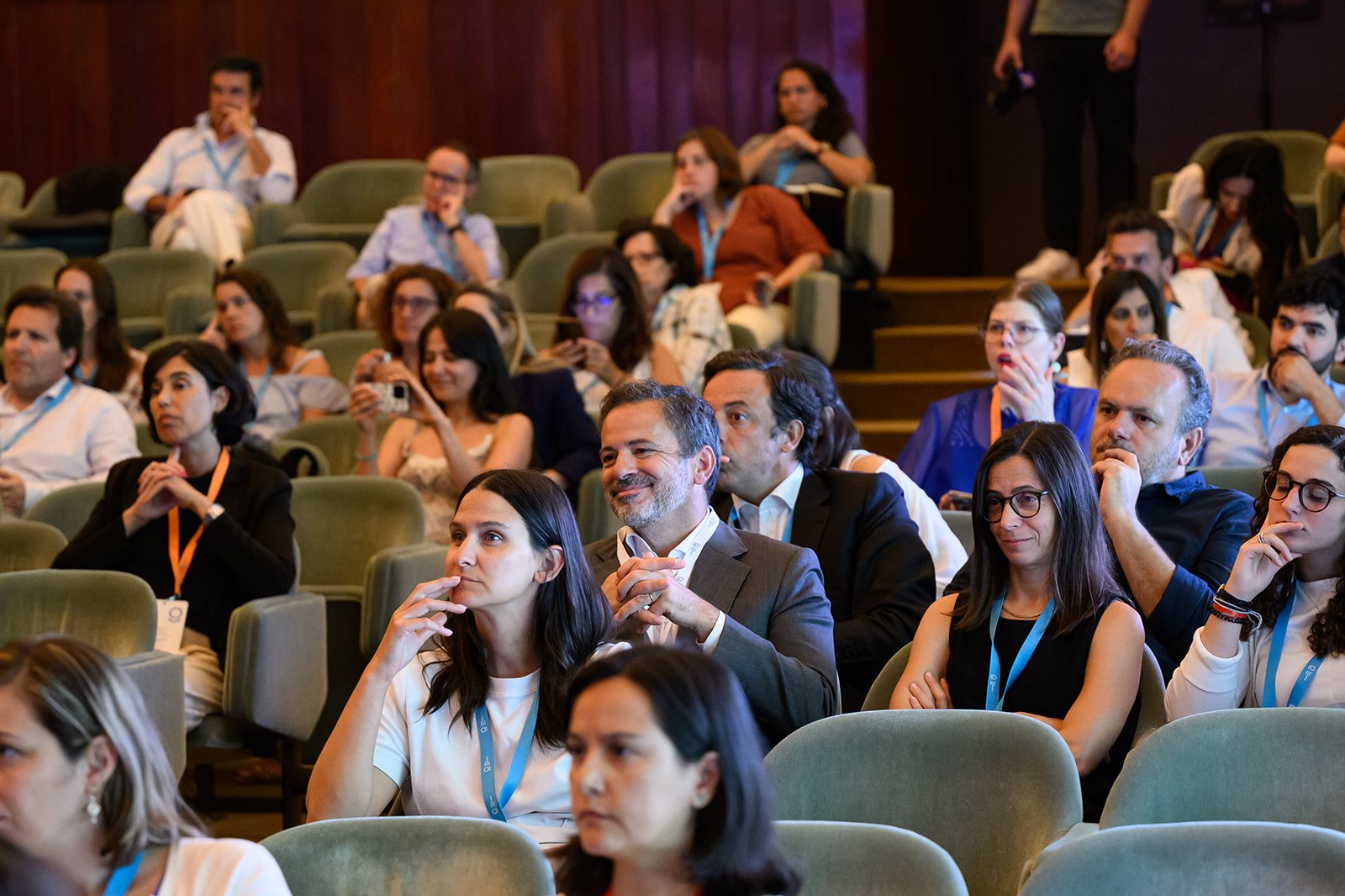Audience seated in a conference hall, attentively listening to a presentation, with some taking notes or photos, wearing conference badges.