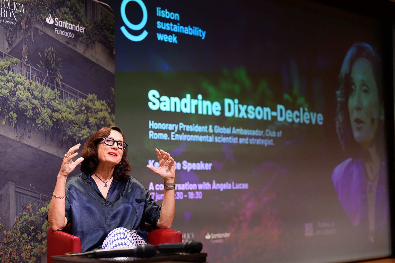 Sandrine Dixson-Declève speaking at Lisbon Sustainability Week, sitting in a red chair, with a large screen behind her displaying event information and a partially visible portrait of her.