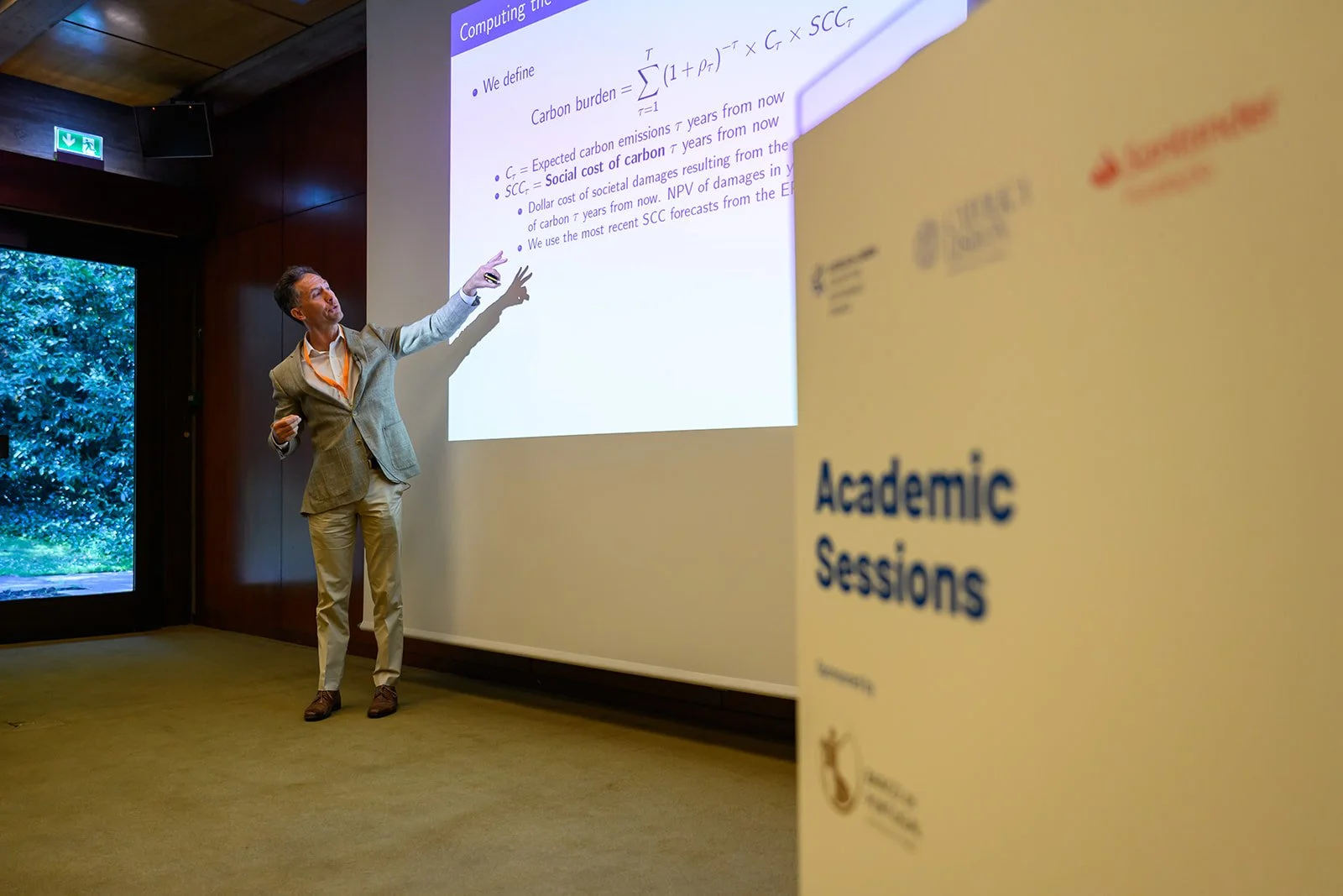 A man giving a presentation at an academic session, pointing at a projected slide on the wall, with a sign in the foreground that reads 'Academic Sessions'.