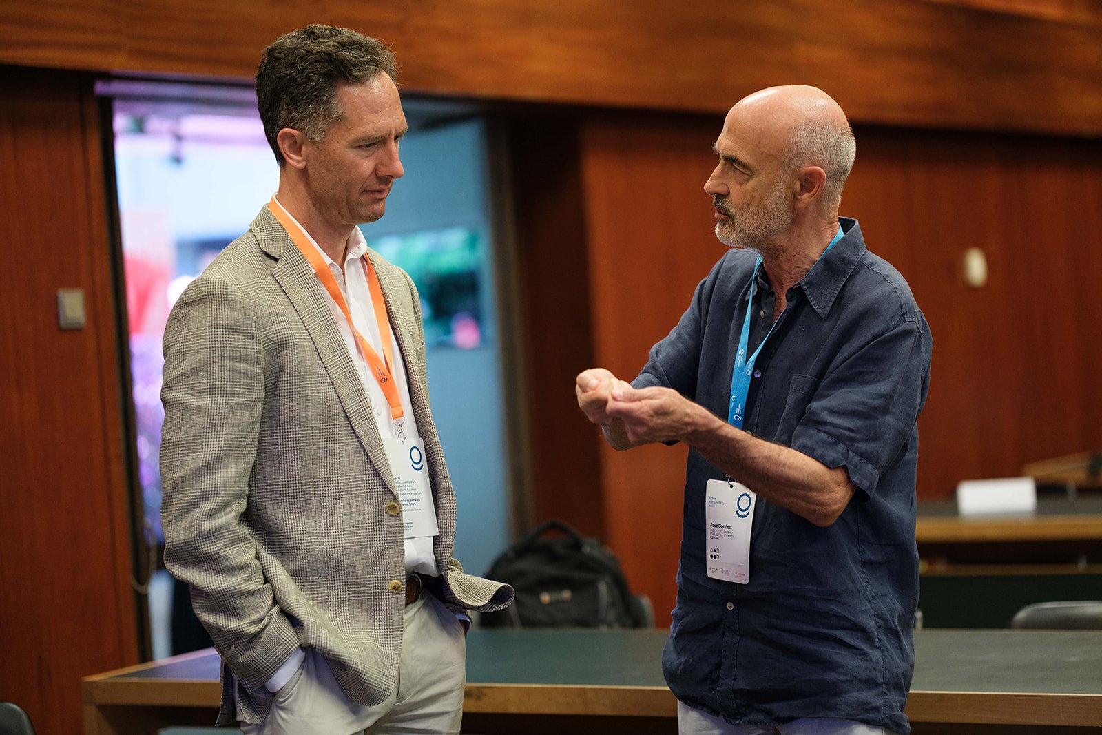 Two men having a conversation at a conference or meeting, wearing conference badges, in a room with wooden panels and a green table.