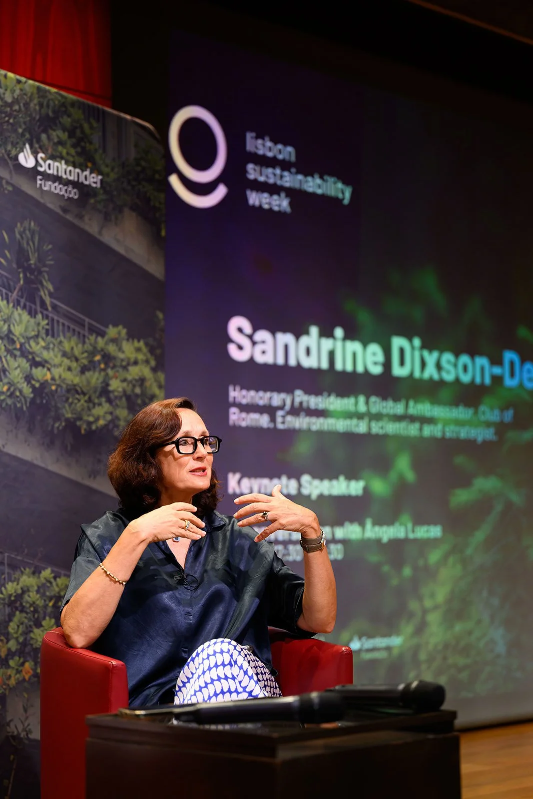 A woman with dark hair, glasses, wearing a dark blouse and patterned pants, sits on a red chair speaking with her hands raised, in front of a large screen displaying text about Sandrine Dixson-Declève at an event for Dublin Sustainability Week.