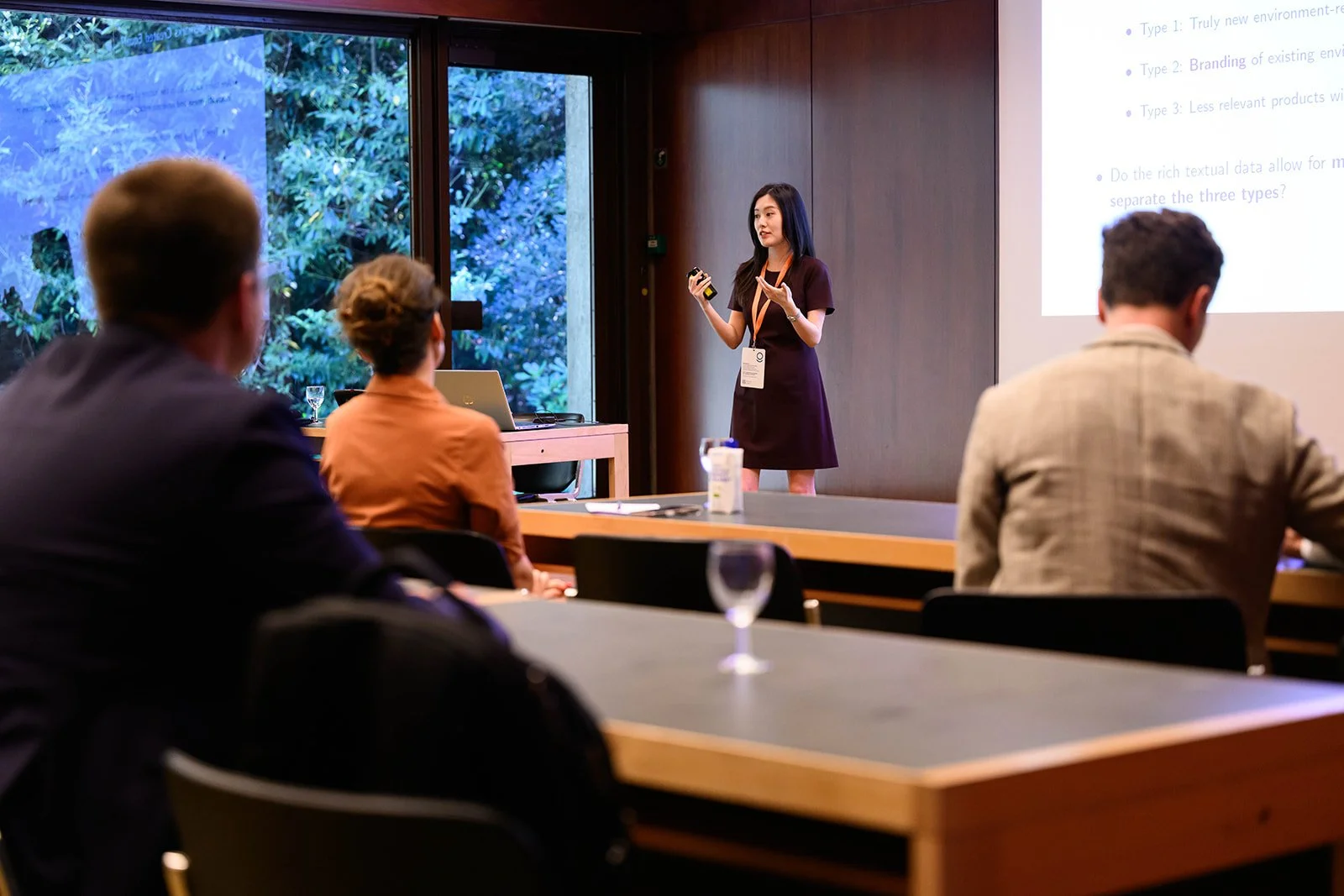 A woman giving a presentation to a group of people seated at tables in a conference room, with a large window showing trees outside.