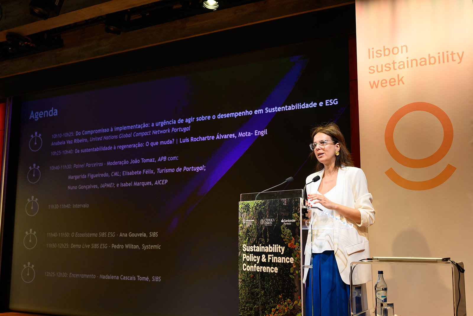 A woman is speaking at a podium during a conference titled 'Sustainability Policy & Finance Conference'. The background features a large screen displaying the agenda of the event in Portuguese and the event branding on the right side reads 'Lisbon Su