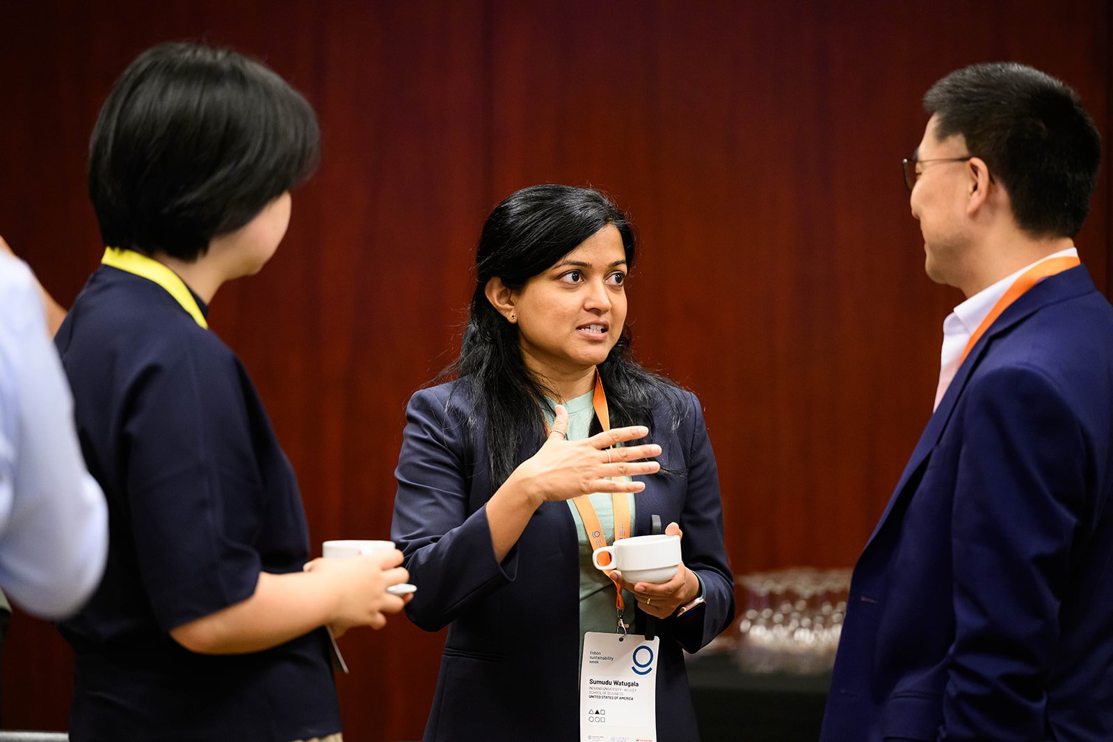 A woman speaking to two other people at a professional event, holding a coffee cup and gesturing with her hand.