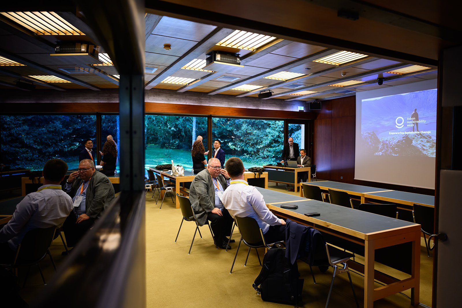 Conference room with several people standing and sitting, large windows overlooking a green outdoor area, a presentation on sustainable change, and a projector screen displaying 'Lisbon Sustainability Week' and 'Empowering Sustainable Change'.