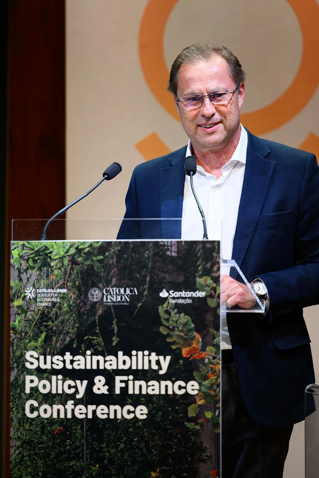 A man in glasses and a blue blazer speaking at a podium during the Sustainability Policy & Finance Conference.