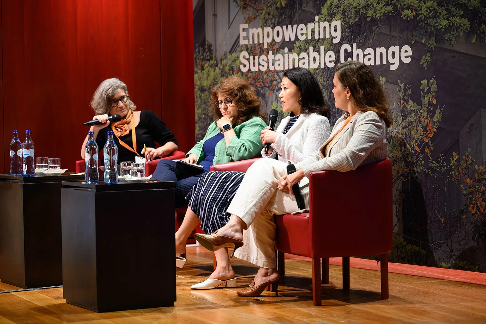 Four women seated on stage at a panel discussion, with the backdrop reading 'Empowering Sustainable Change'. One woman is speaking into a microphone, others are listening or taking notes. There are water bottles and glasses on small tables beside the
