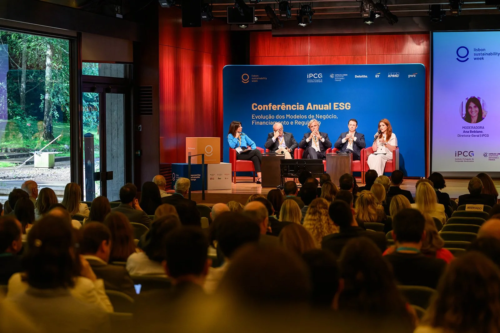 A panel discussion at the Lisbon Sustainability Week conference with five speakers on stage, a large blue banner behind them, and a seated audience watching inside a conference hall.