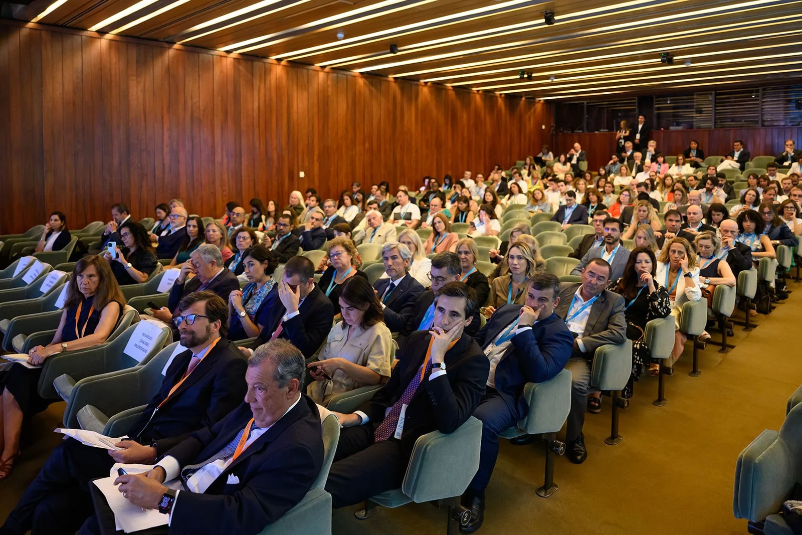 A large audience seated in a conference room with wooden walls and green chairs, attending a formal event.