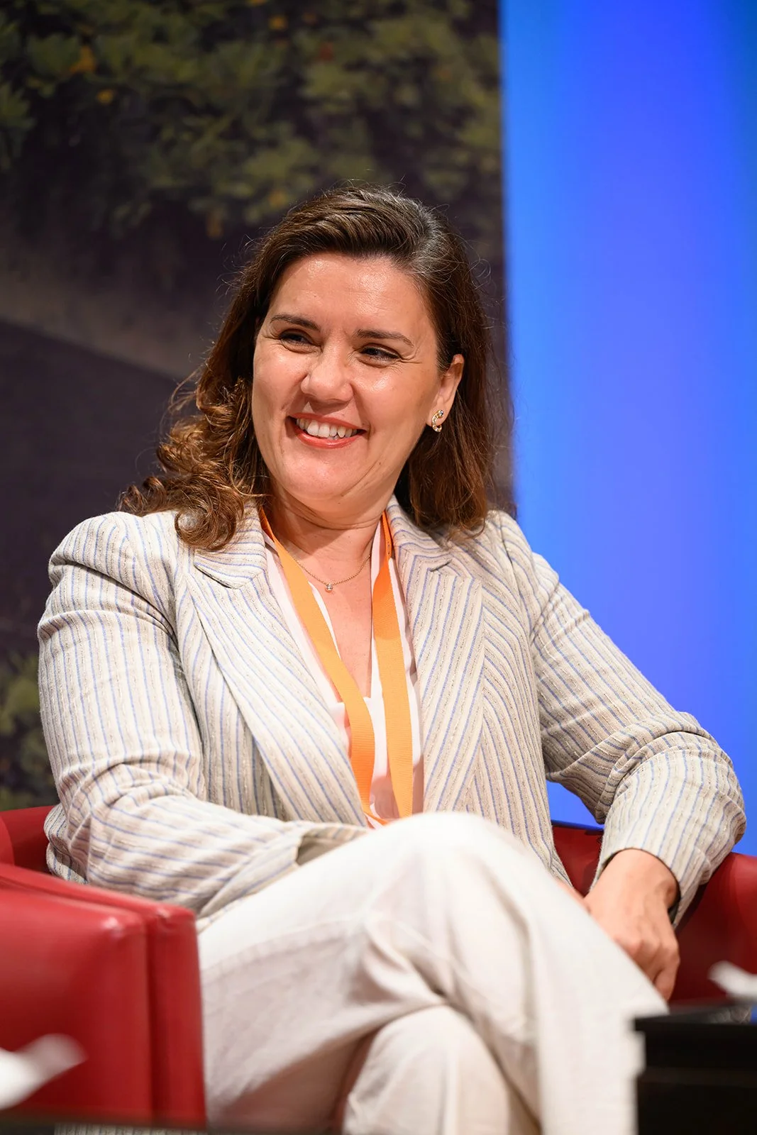 Woman with brown hair smiling, sitting on a red chair, wearing a striped blazer and a lanyard, in a professional setting.