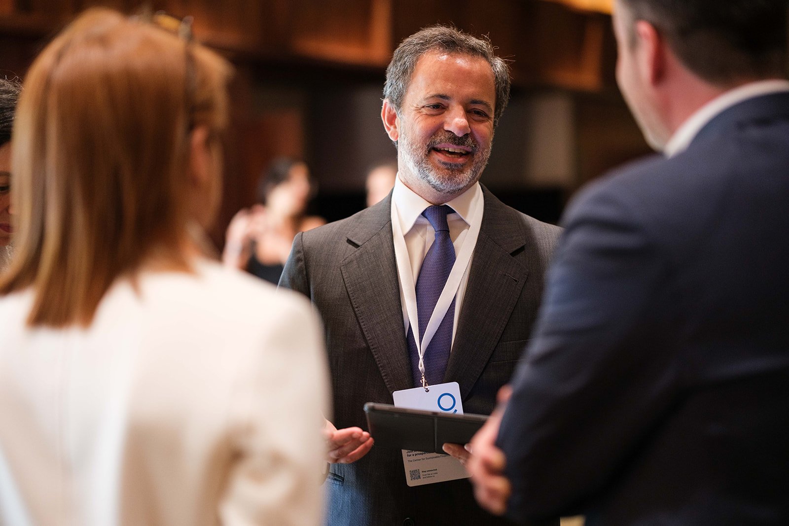Group of people at a professional networking event, woman with red hair and man are talking to a smiling man in a suit with a name badge, in a warmly lit room.