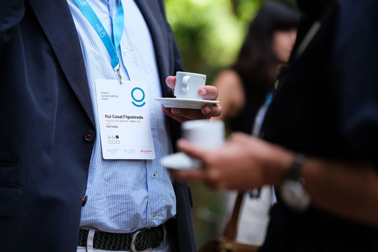 Person wearing a conference badge with the name Rui Casal Figueiredo, holding a white cup and saucer, at an outdoor event with greenery in the background.