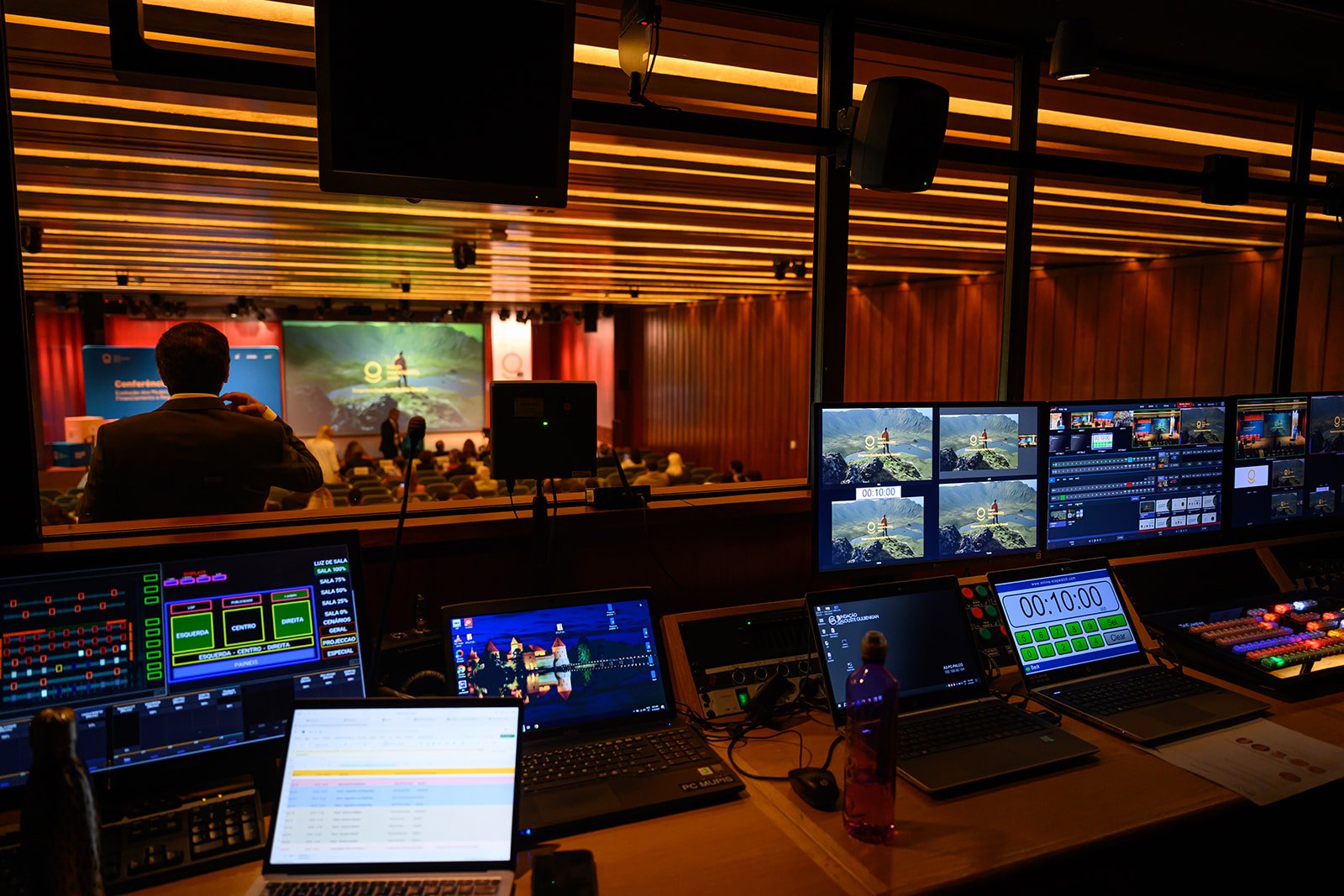 View of a conference or presentation event from control room with multiple monitors displaying live feed and technical controls; audience seated facing stage with large screen, presenter, and red curtains, illuminated by warm lighting
