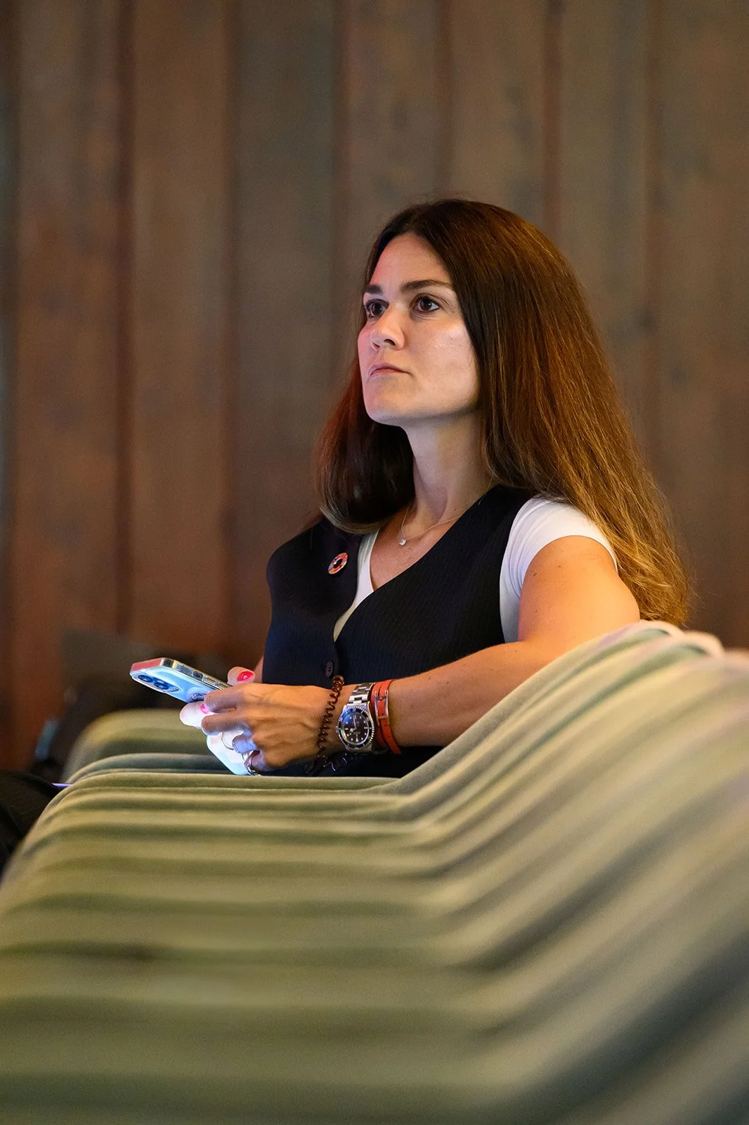 A woman with long brown hair sits on a striped green and beige couch, holding a cell phone, wearing a white shirt with a black vest, in a room with wooden wall paneling.