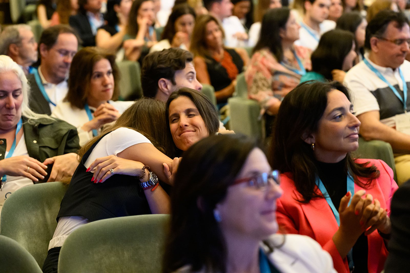 A group of people sitting in an auditorium or conference room, with two women in the center hugging and one smiling woman hugging her. The audience appears emotional, some with eyes closed or looking down, in a professional or formal setting.