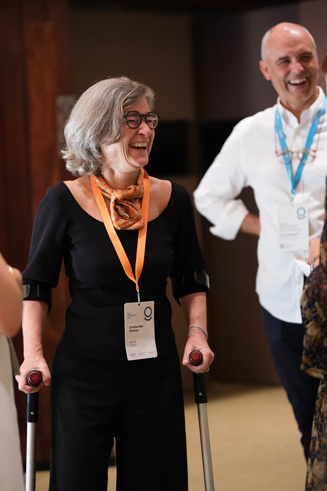 A woman with gray hair, glasses, and a patterned scarf around her neck, smiling and laughing while holding crutches, standing next to a man with a bald head, white shirt, and blue lanyard, also smiling and standing with hands on hips, in a conference