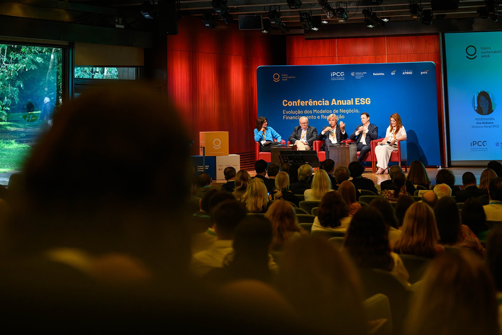 A conference panel discussion with five seated panelists on stage, with a blue backdrop displaying the event title in Portuguese, and a large audience seated facing the stage.