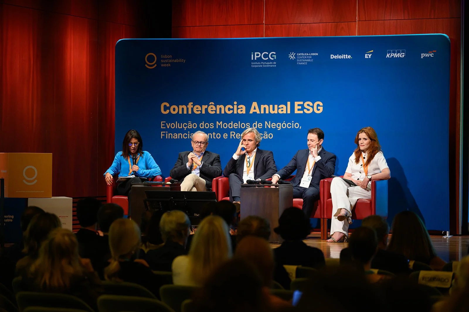 Panel of five speakers seated on stage at the Lisbon Sustainability Week conference. The background displays a blue banner with the conference title, sponsors, and logos. Audience members seated in front are visible.