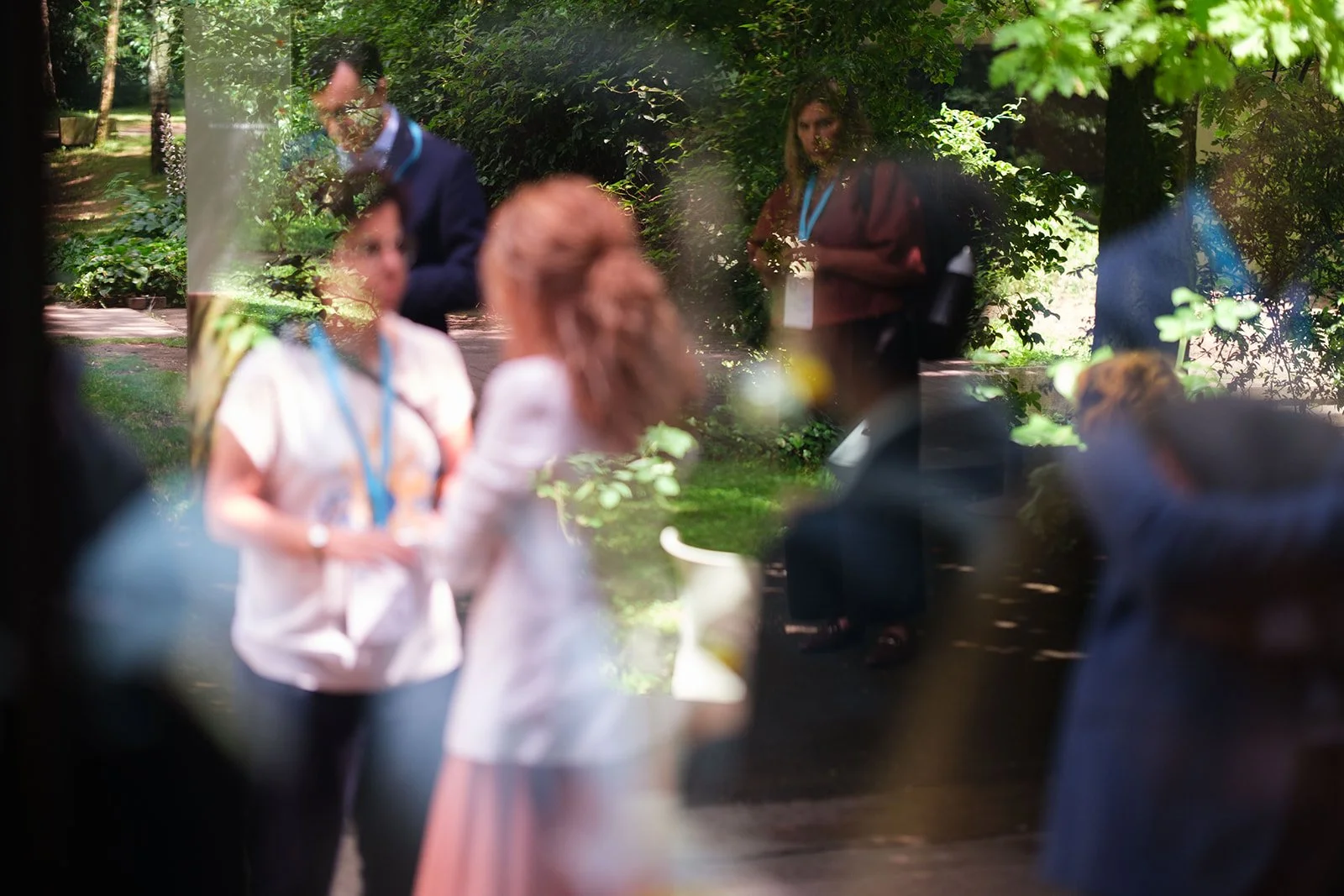 People seen through a glass window during an outdoor event on a sunny day with green trees and foliage in the background.