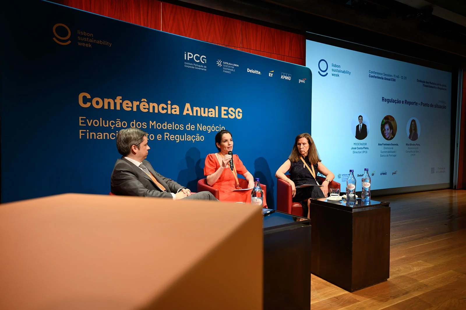 Three people seated on stage at a conference, with a large blue backdrop displaying event details and logos. The backdrop reads 'Conferência Anual ESG' and describes the event about the evolution of financial business models and regulation. The woman