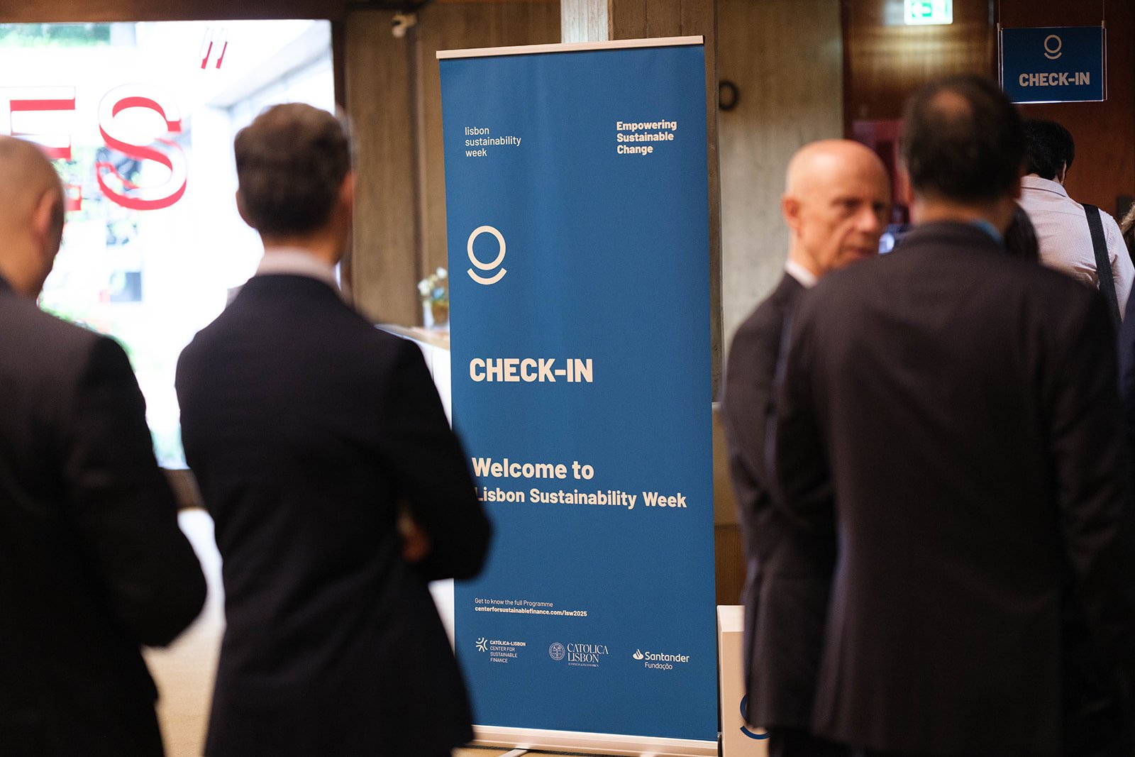 People gather near a blue sign that reads 'CHECK-IN Welcome to Lisbon Sustainability Week' at a conference event.