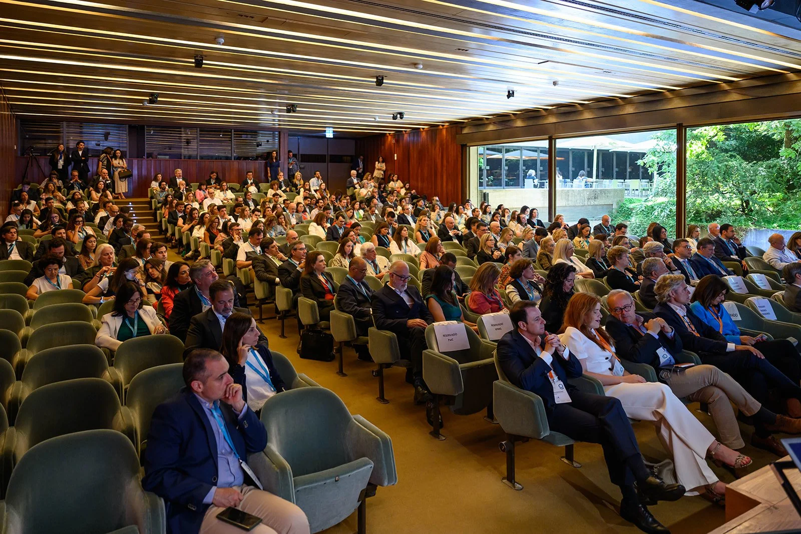 Large conference room filled with seated attendees during a presentation, with a view of outside greenery through large glass windows.