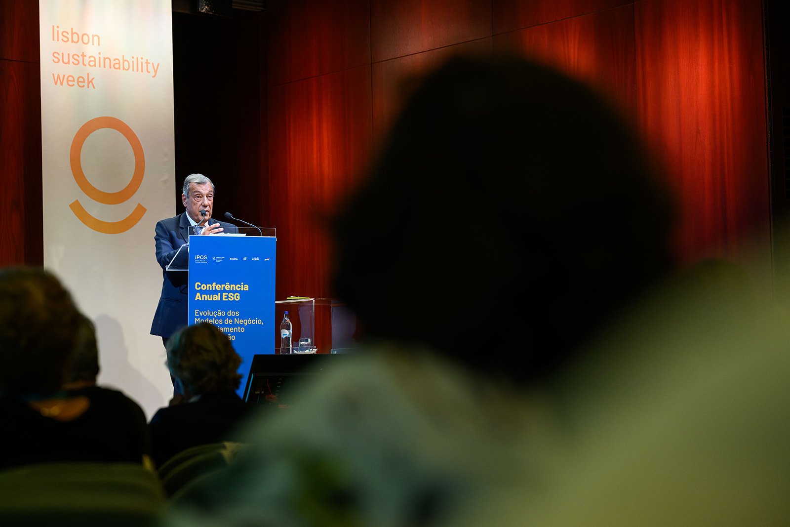 A man in a suit standing at a podium giving a speech during a conference on sustainability, with an audience and a banner in the background.